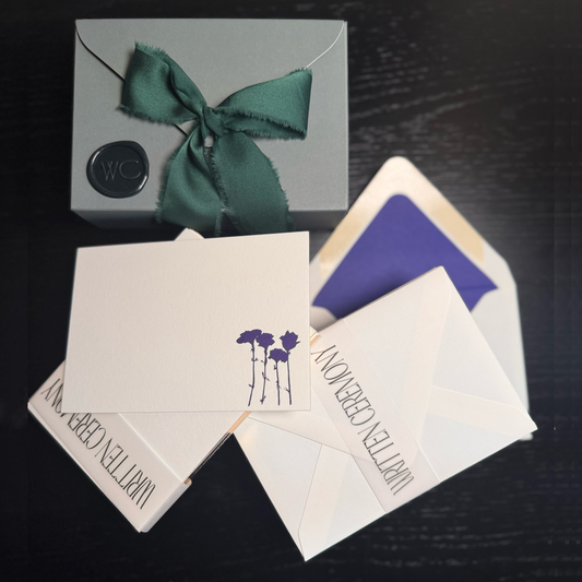 Set of ten luxury stationery note cards with gilded gold edges with a cobalt design of four flowers with cheeky women hanging as leaves. A  green envelope shaped gift box with wax seal is enclosed by a dark silk green ribbon.