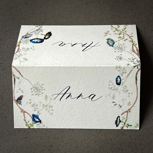 Elegant folded place card with delicate blue floral and butterfly border design, featuring handwritten name "Anna" in modern script — perfect for chic, modern tablescapes and sophisticated hostess dinners by Written Ceremony.